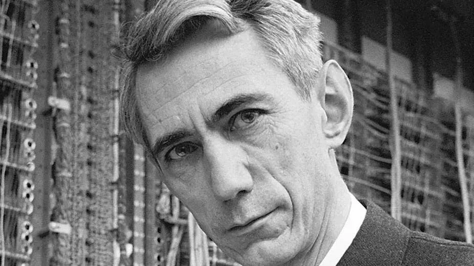 A black-and-white portrait of Claude Shannon in front of telephone switching equipment.